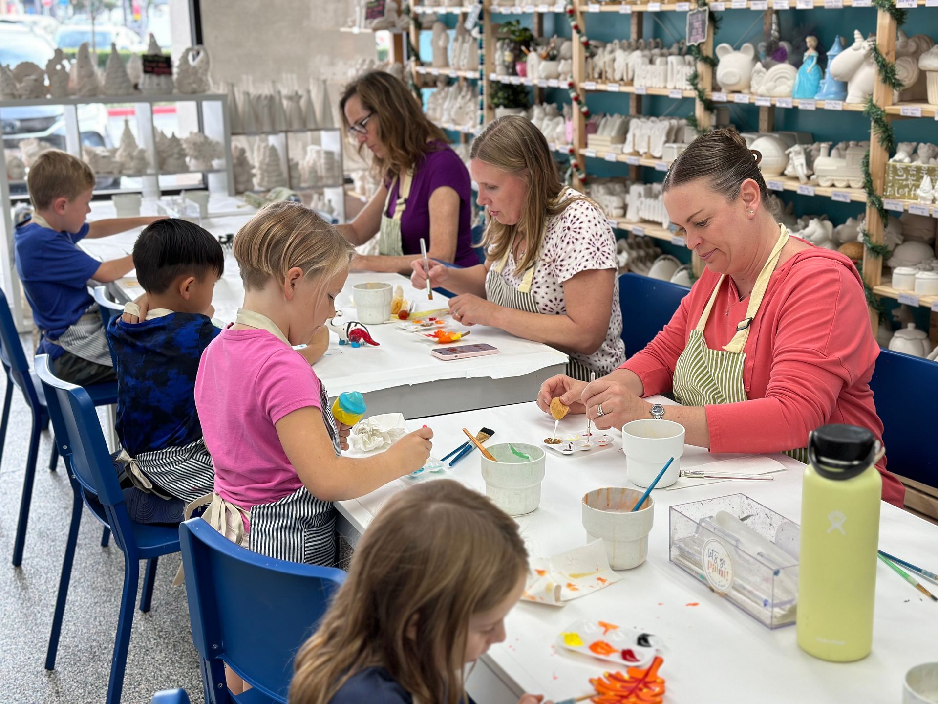 A group of people are sitting at a table in a pottery studio. They can be seen painting ceramics.