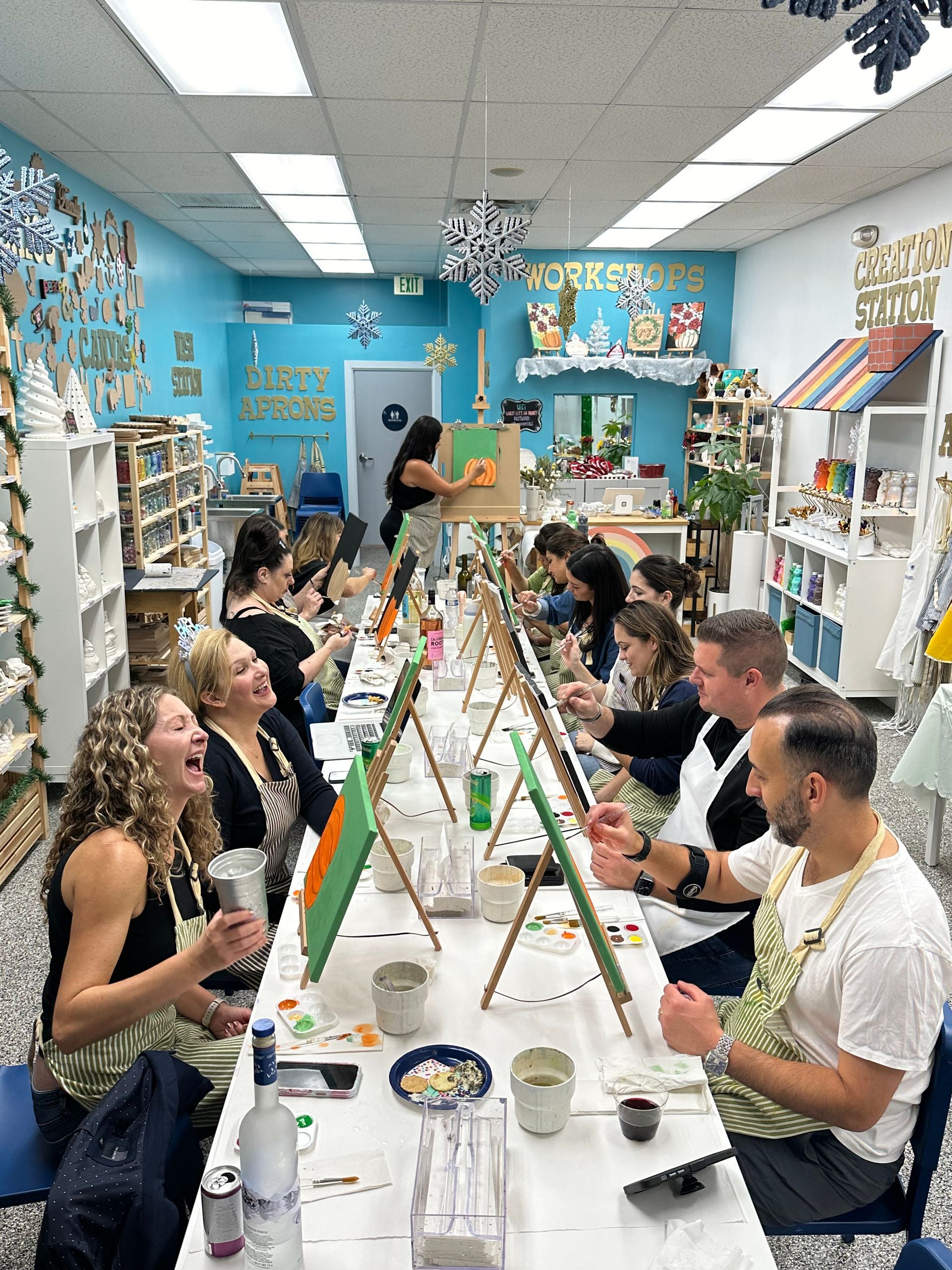 A group of people are sitting at a long table painting. There is a teacher at the end of the table. The group is learning how to paint a fall scenery filled with pumpkins.