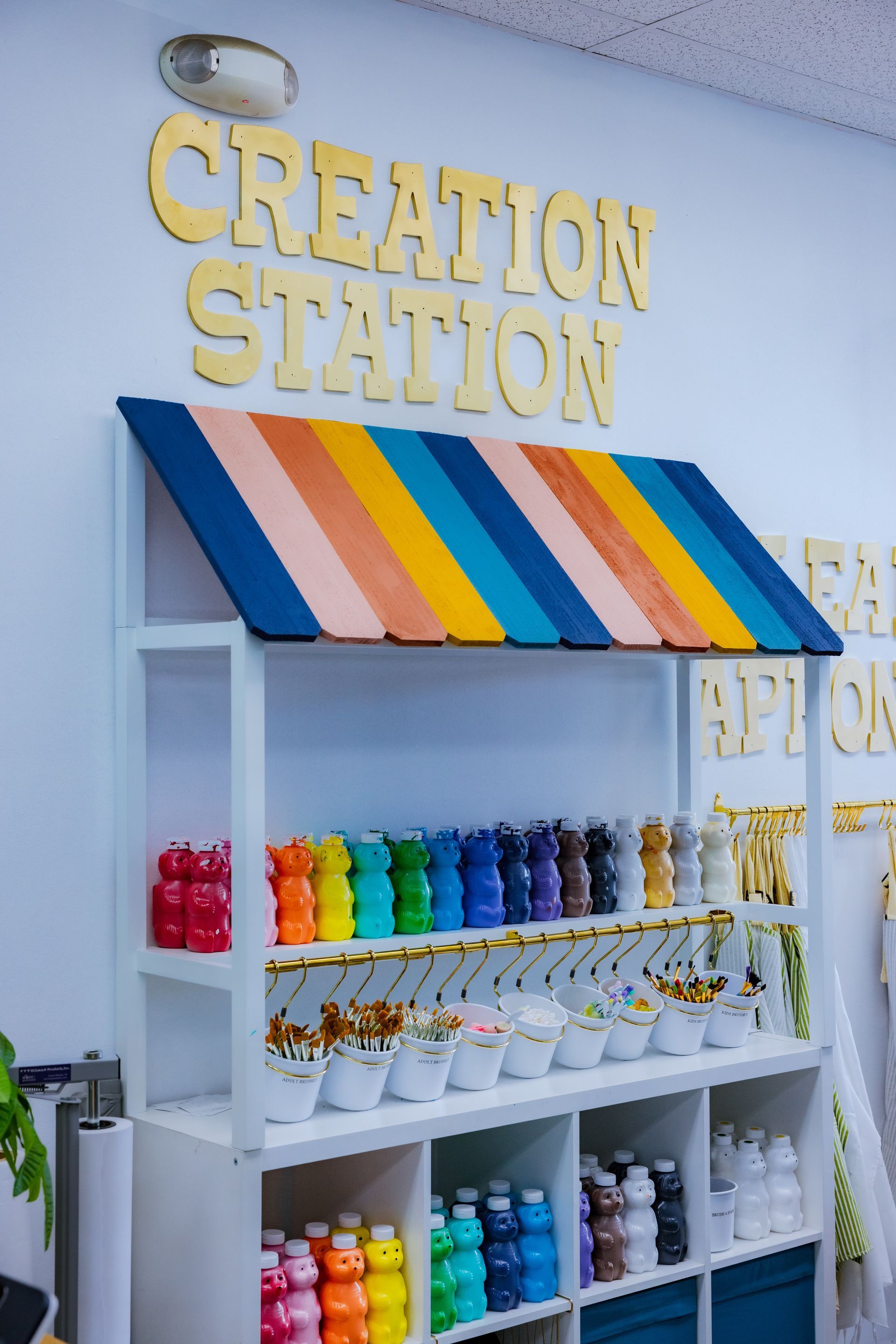 A colorful stand on a wall displays bottles of paint. These bottles of paint are in a honey-bear shape. On top it is labeled, 