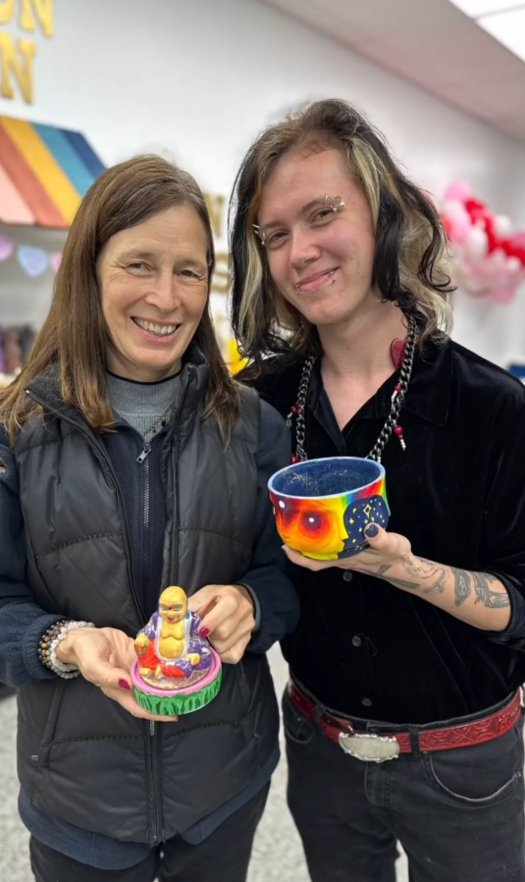 Two women are standing next to each other holding ceramic creations. One is holding a Buddha statue, another holding a colorful bowl.