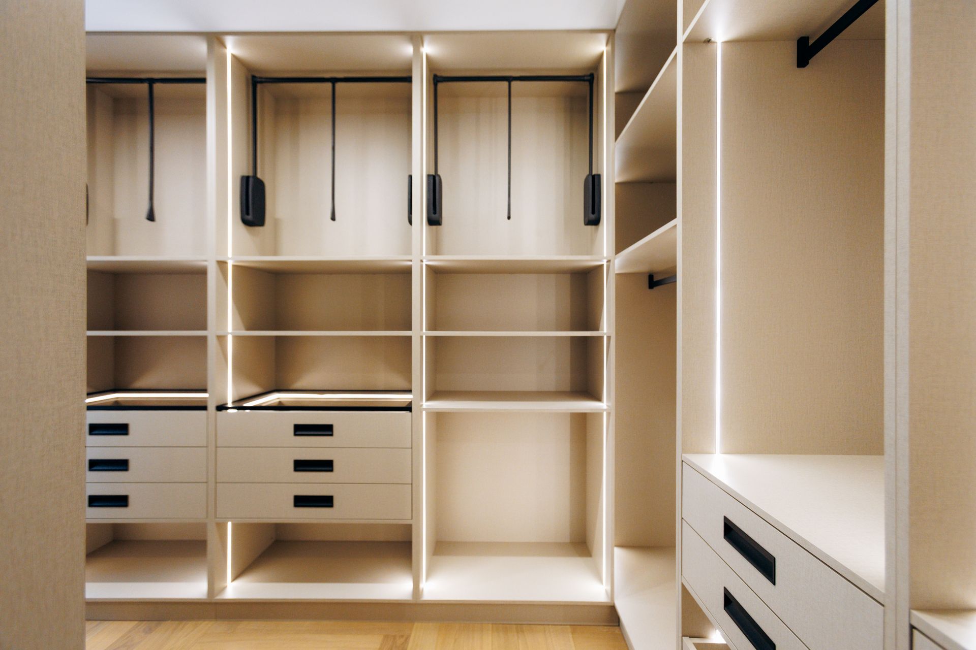 Empty walk-in closet with beige shelves, drawers, and hanging rods under recessed lighting