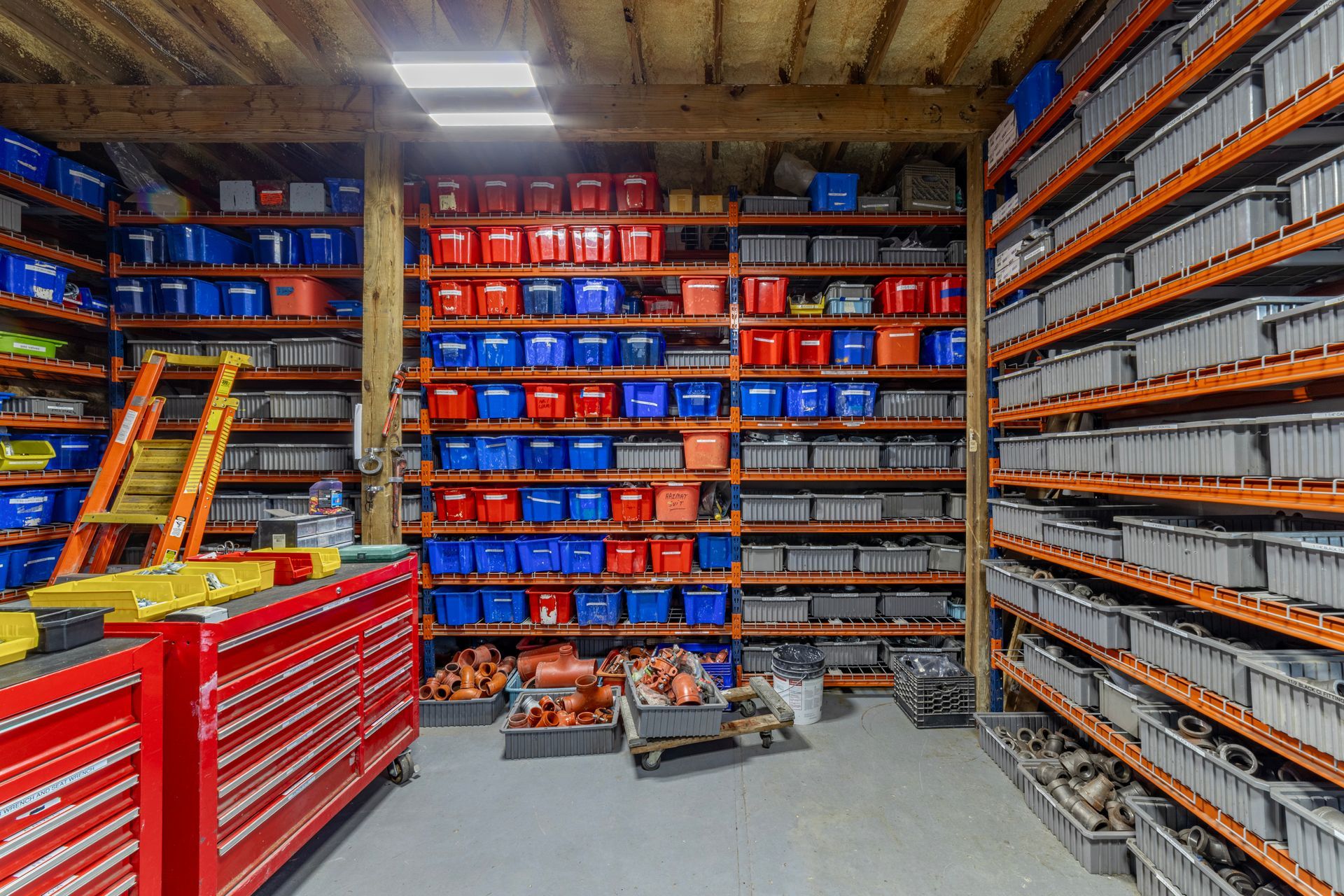 Storage warehouse aisle with red tool chests, shelves of blue and orange bins, and stacked parts.