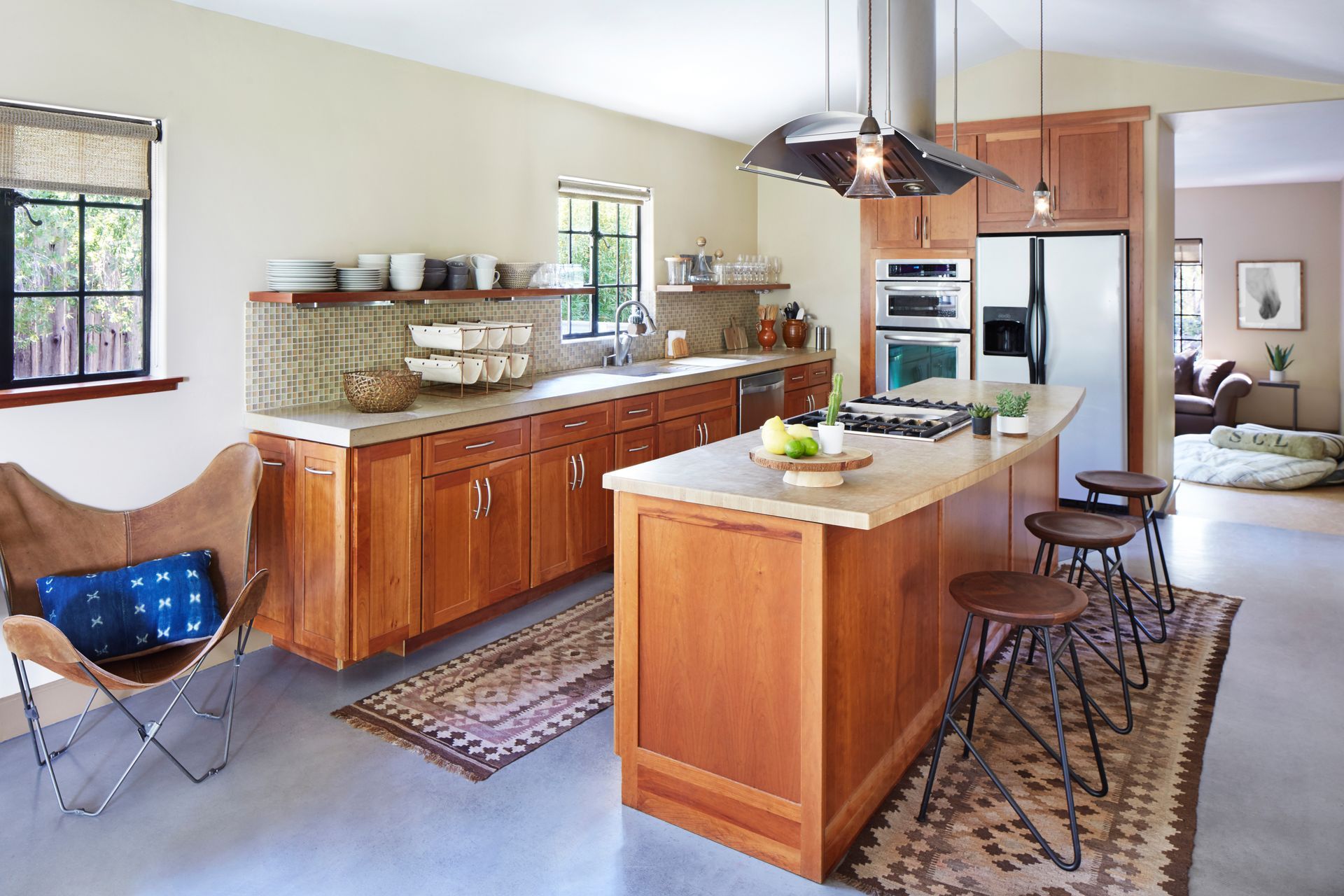 Warm kitchen interior with wood cabinets, island, stools, rugs, and a blue folding chair by the window