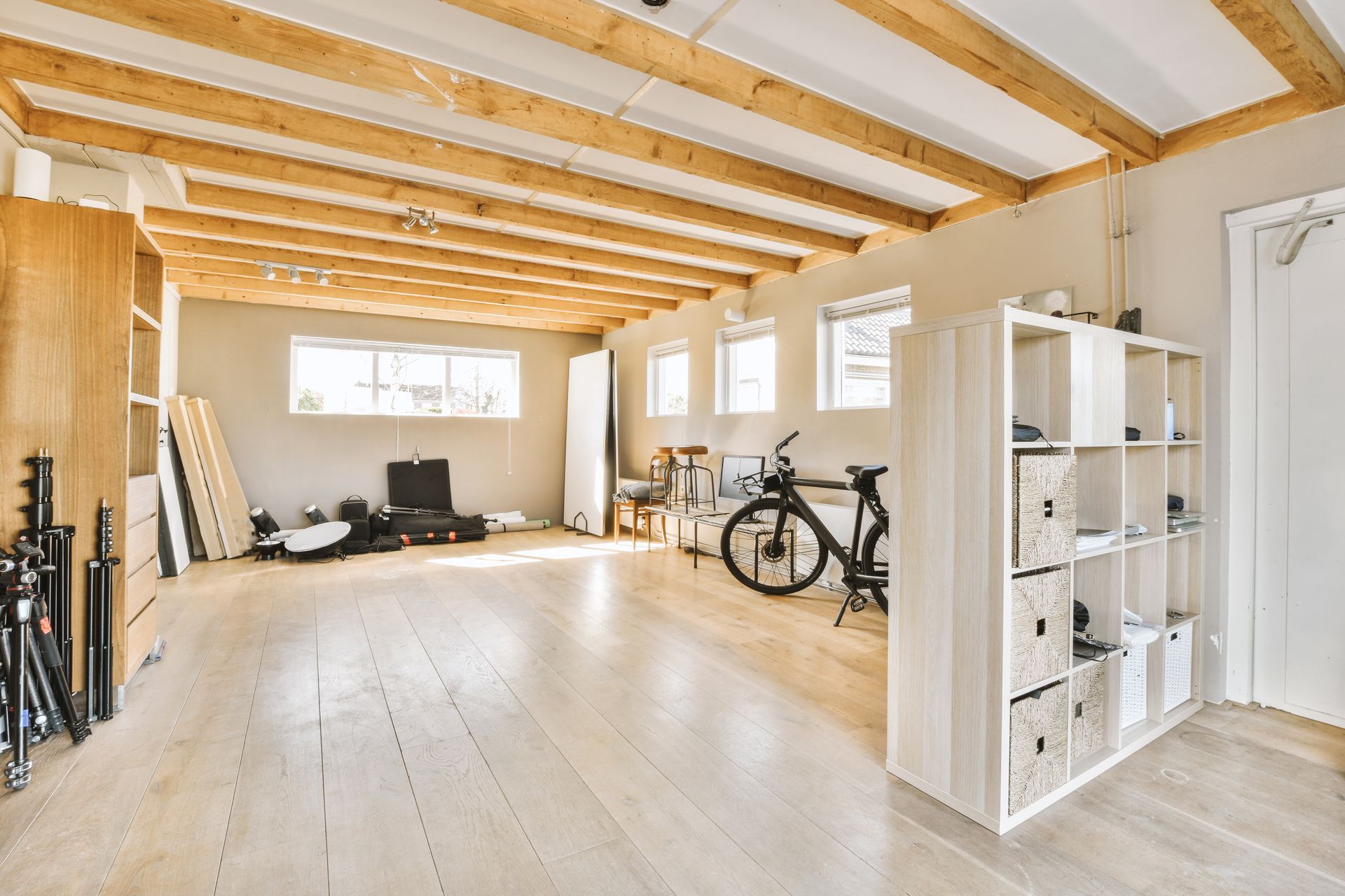 Bright, unfinished loft room with exposed beams, bikes, shelving, and large windows