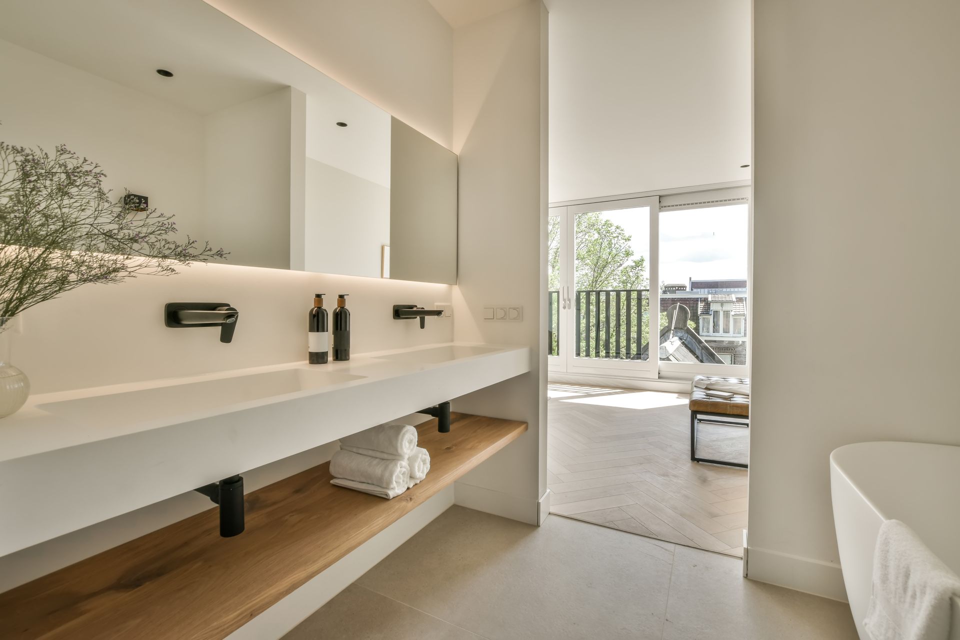 Modern bathroom with floating double sink, beige walls, and open balcony view beyond a bedroom doorway