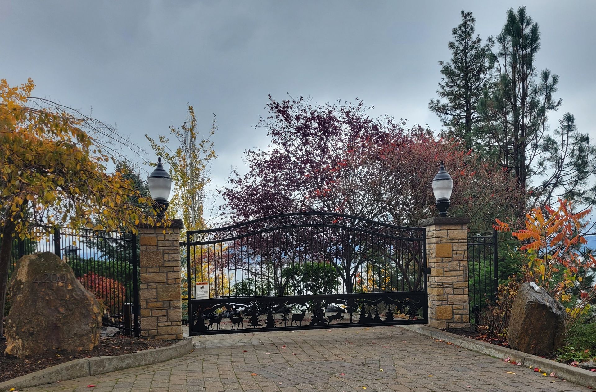 Ornate black gate with stone pillars and brick driveway, trees with autumn colors in background.