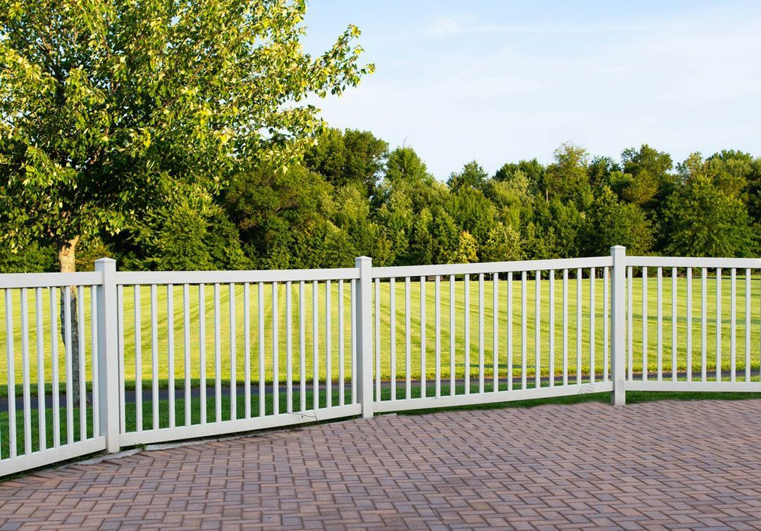 A white fence surrounds a brick driveway with trees in the background.