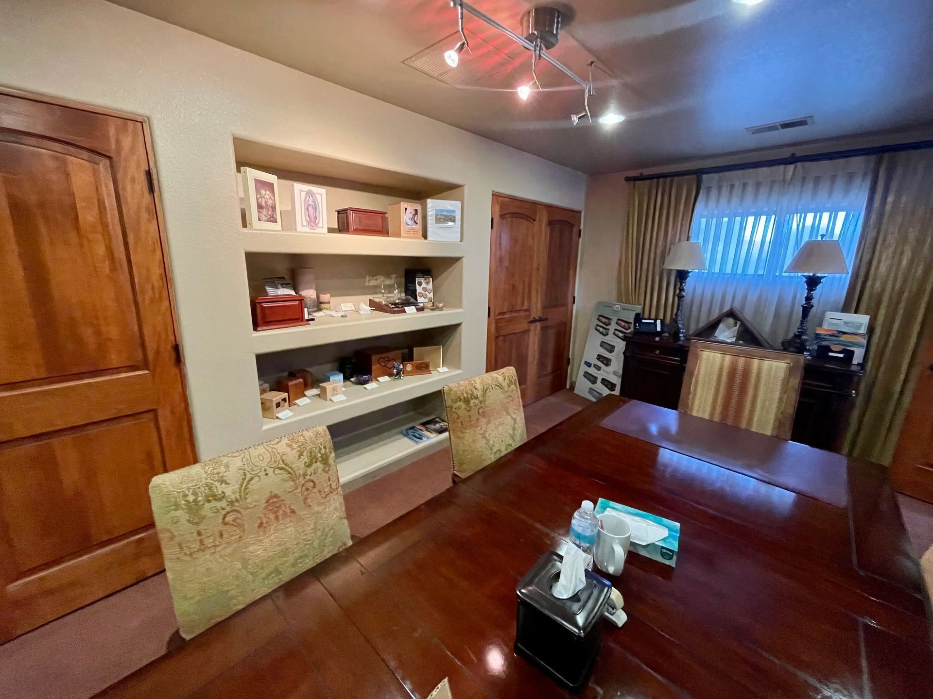 Wooden-paneled office with a built-in shelf displaying items, a long table with chairs, and a window with curtains.