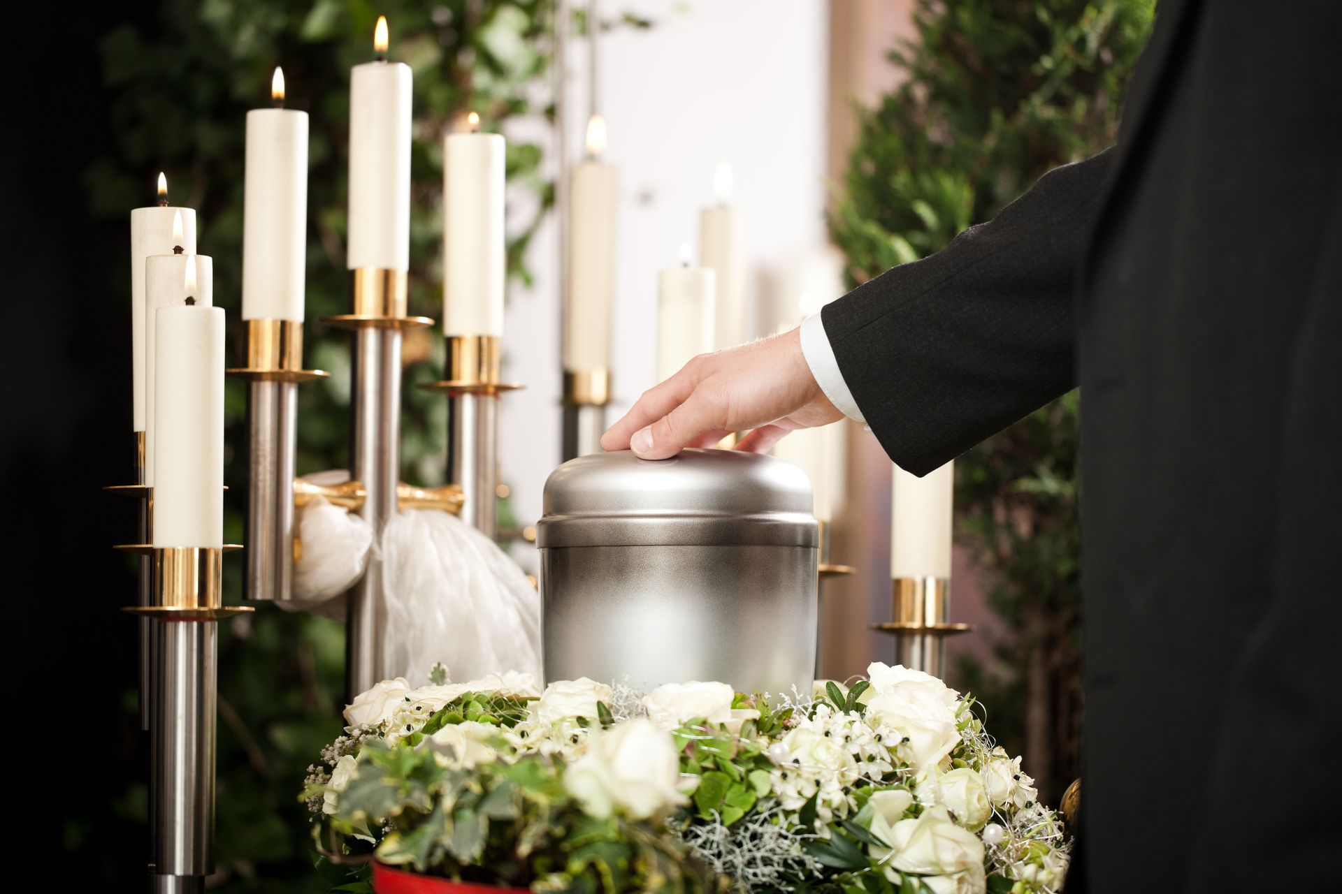 Person's hand on a silver urn, surrounded by flowers and candles at a memorial service.