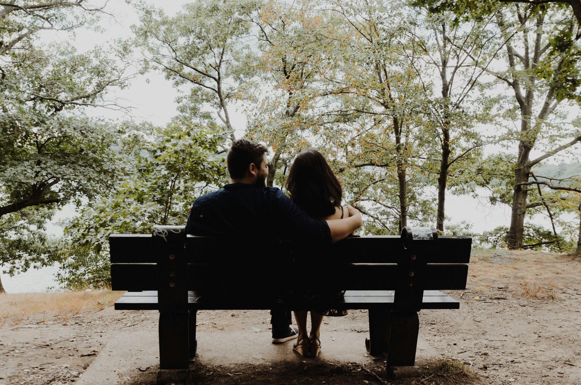 Couple seated on park bench, facing water, trees in the background.