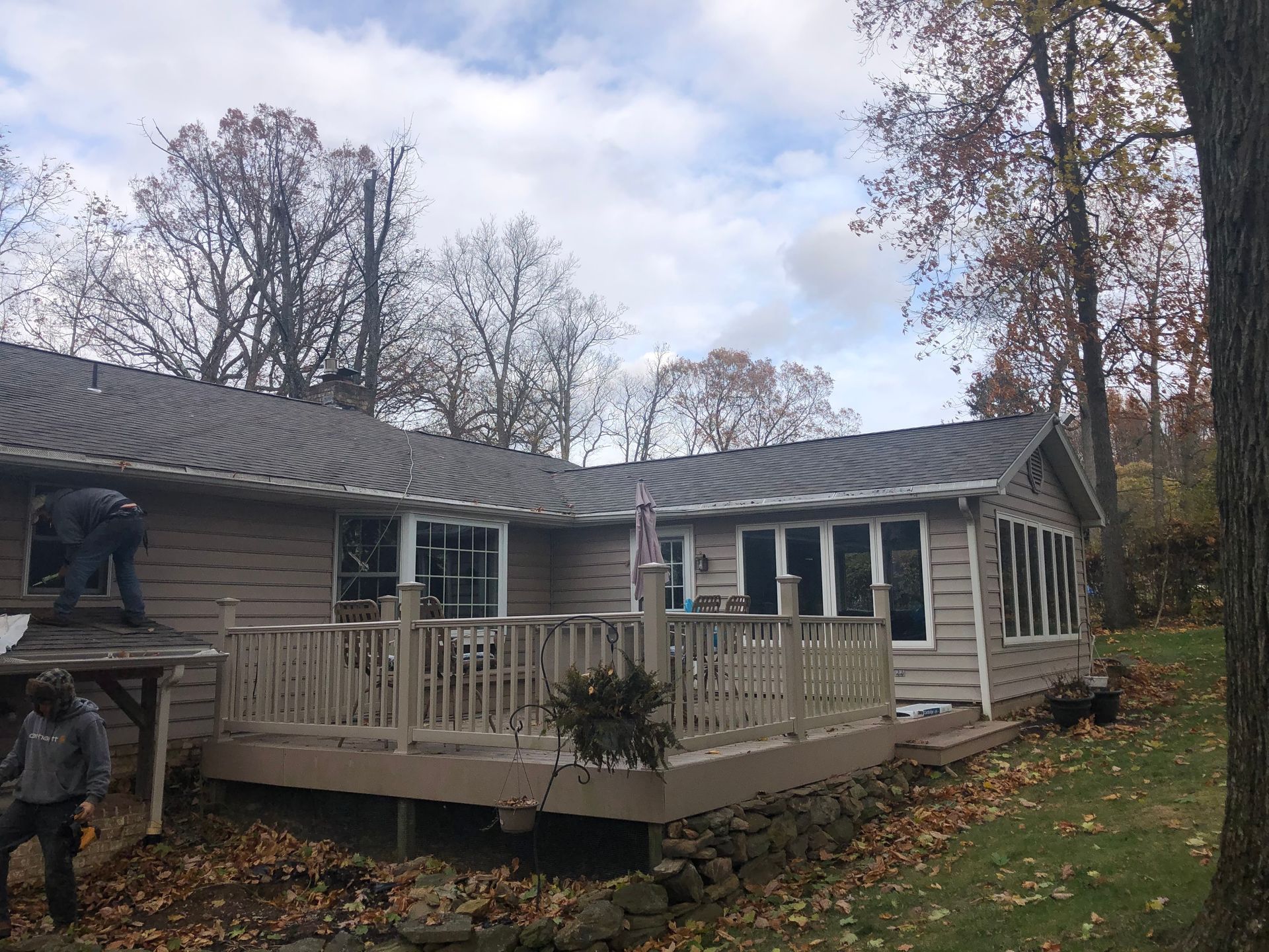 A house with a deck; two people work on the roof. Brown siding and a rock wall in a yard with trees.