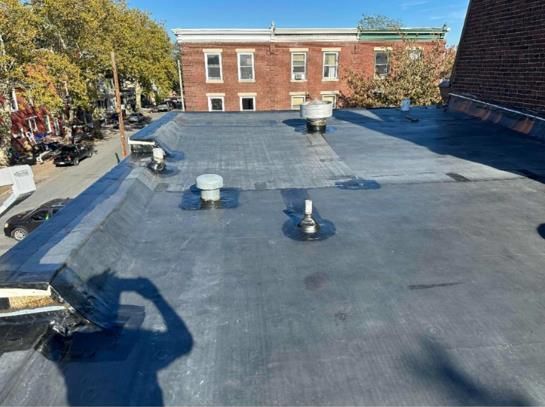 A flat, dark rooftop with several vents. A brick building is in the background. The shadow of the photographer is visible.