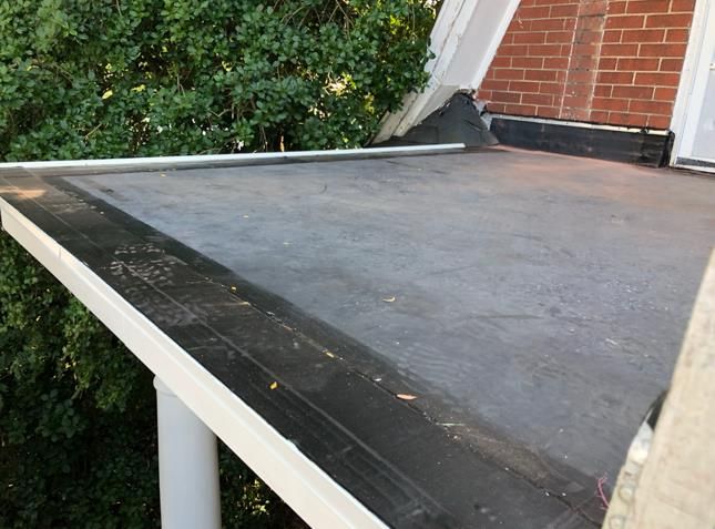 Flat, black roof with white trim, near a brick chimney and lush green trees.