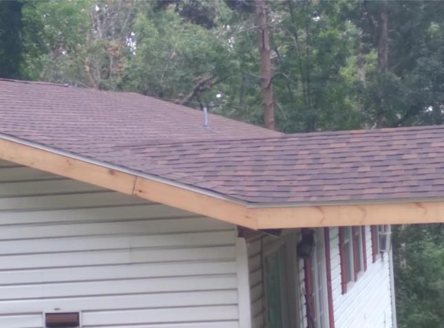 Brown shingle roof with light-colored wooden trim on a house with white siding and a green background of trees.