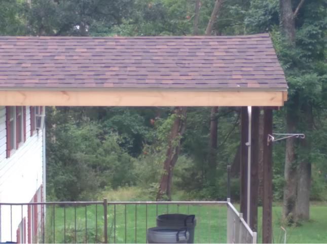 Brown-shingled roof extending over a deck with a dark metal railing, viewed against a background of trees.