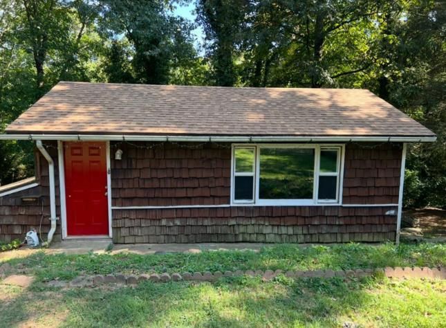 Small, brown-shingled building with a red door and window, set in front of trees, with grass in the foreground.