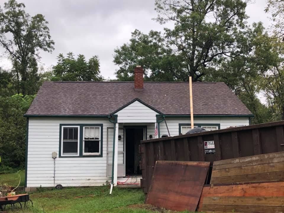 White house with brown roof, green trim, and open door; dumpster and wood planks in front.
