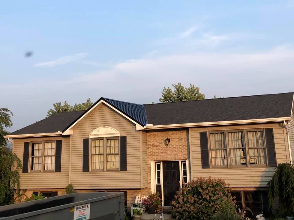 House with new dark roof, beige siding, black shutters. Blue sky in background.
