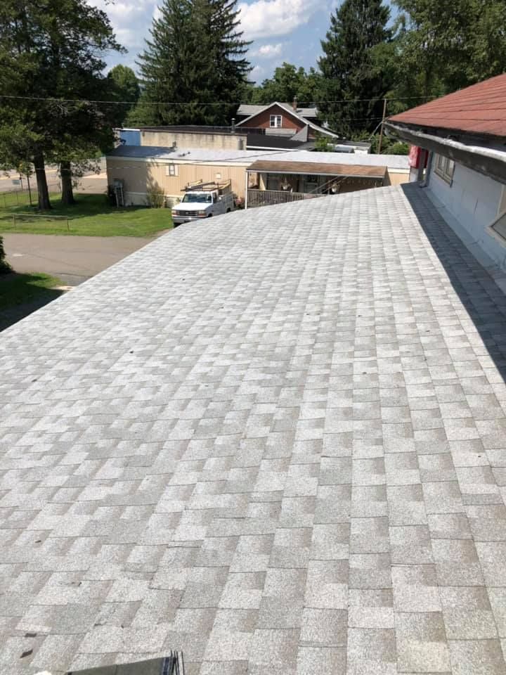 Gray asphalt shingle roof on a building, seen from above, on a sunny day. Background includes trees and other buildings.