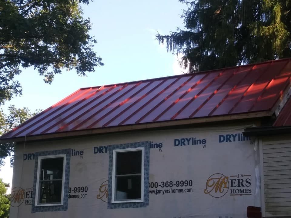 Red metal roof on a house under construction; two windows below, blue sky.