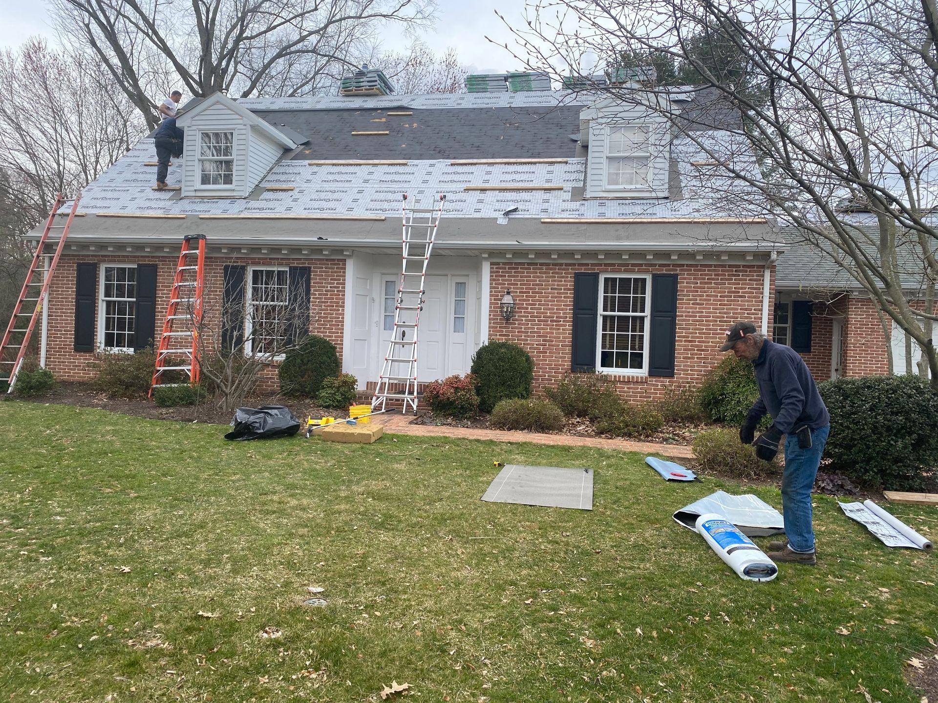 Roofers working on a brick house roof with ladders and materials on the lawn.