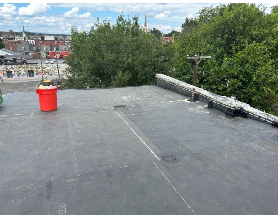 Flat black roof with an orange barrel, surrounded by trees and buildings. Blue sky.
