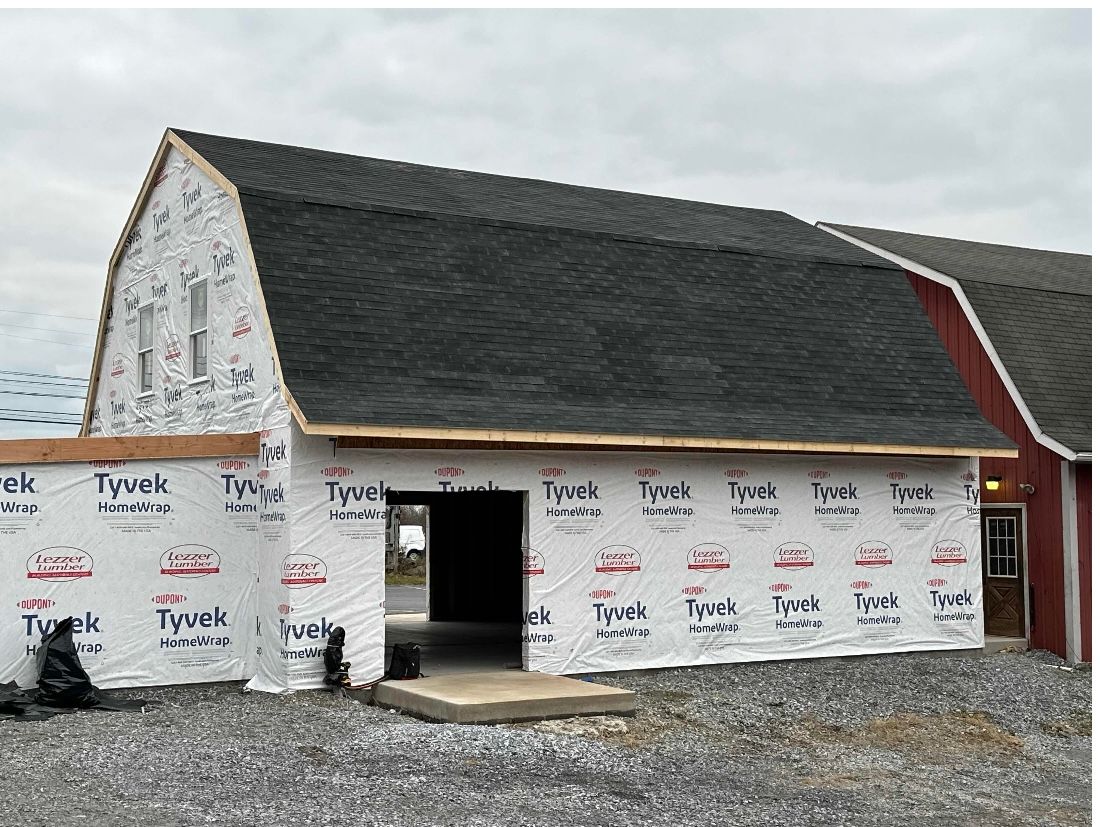 Red barn under construction, with dark roof, Tyvek wrap, and gravel ground.