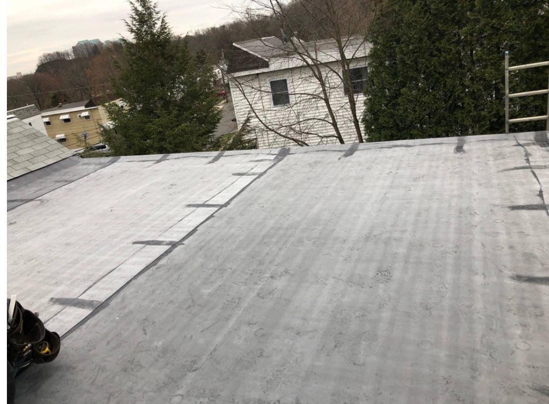 Gray flat roof with dark gray strips, ladder in the background, trees, and white building.