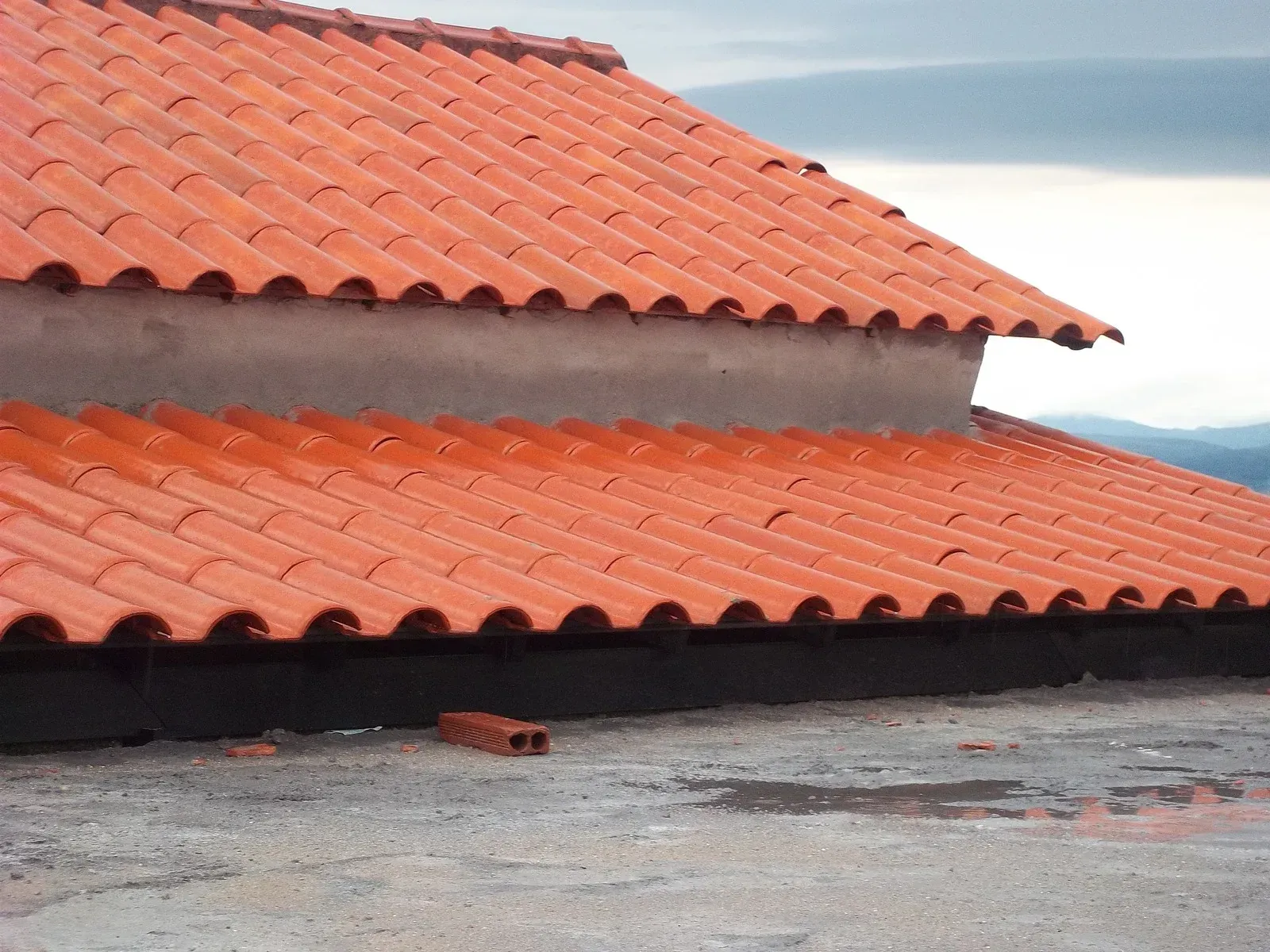 Red tiled roof on a building, with a cloudy sky in the background. A brick sits on the flat surface in the foreground.