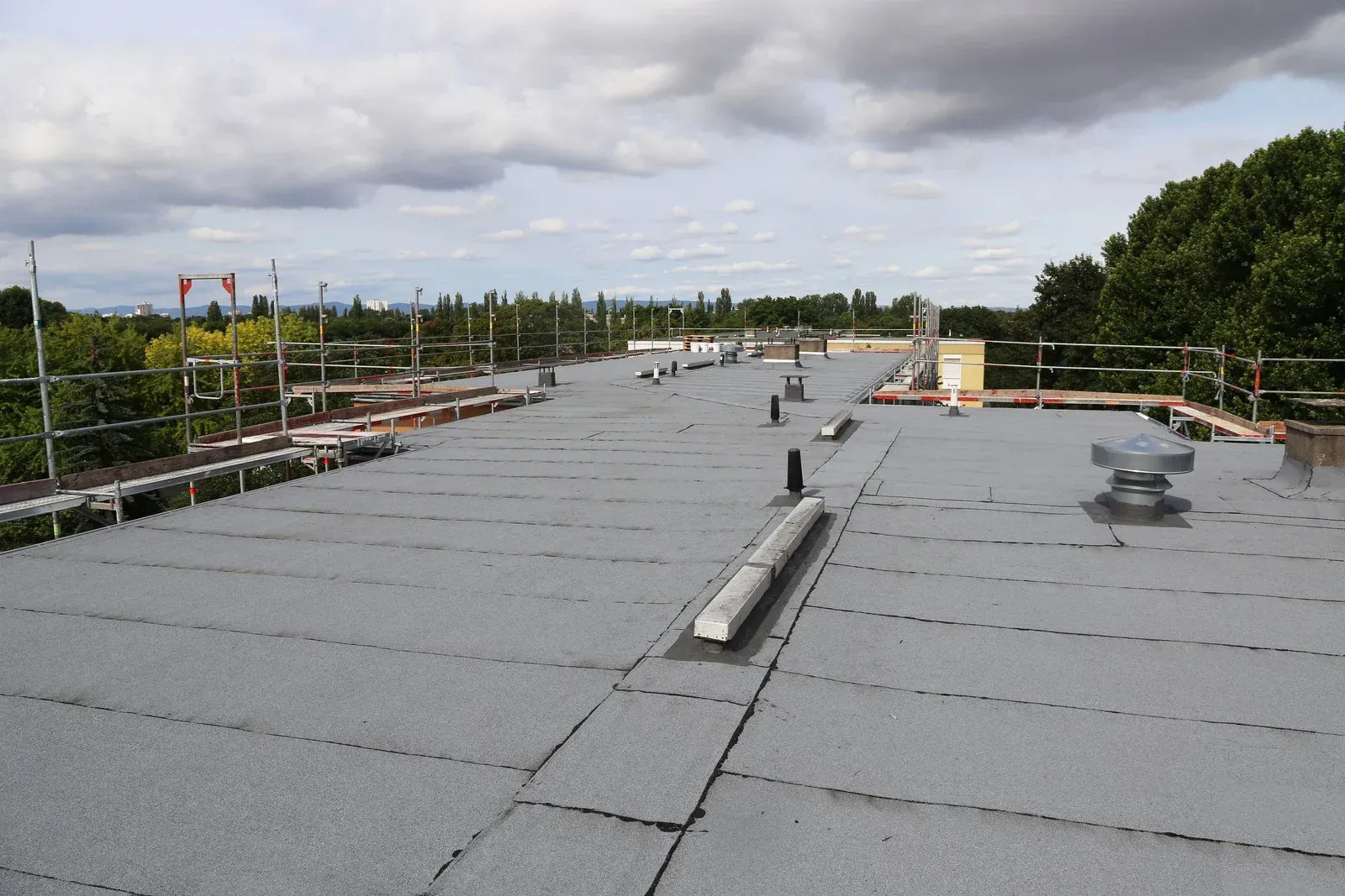 Flat, gray commercial roof with vents and pipes, surrounded by scaffolding and trees under a cloudy sky.