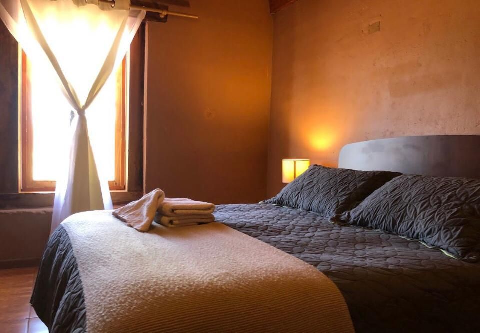 Dormitorio con cama cubierta con una manta acolchada marrón. Una ventana con cortinas transparentes proporciona luz.