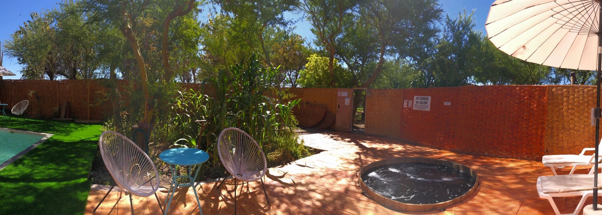 Patio con sillas, mesa y piscina. Árboles rodean una valla roja.