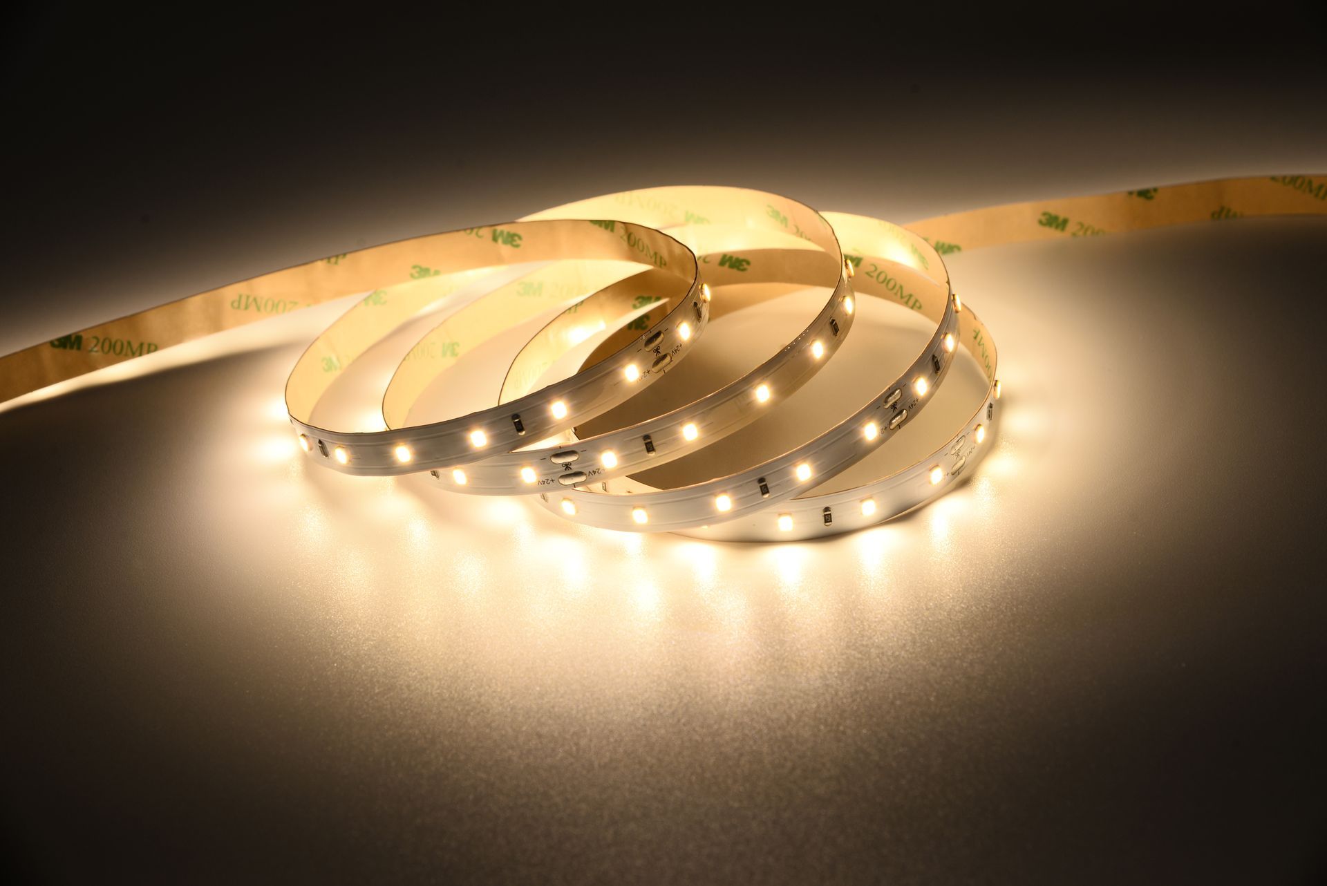 Coiled LED light strip emitting warm white light. The strip has a self-adhesive backing.