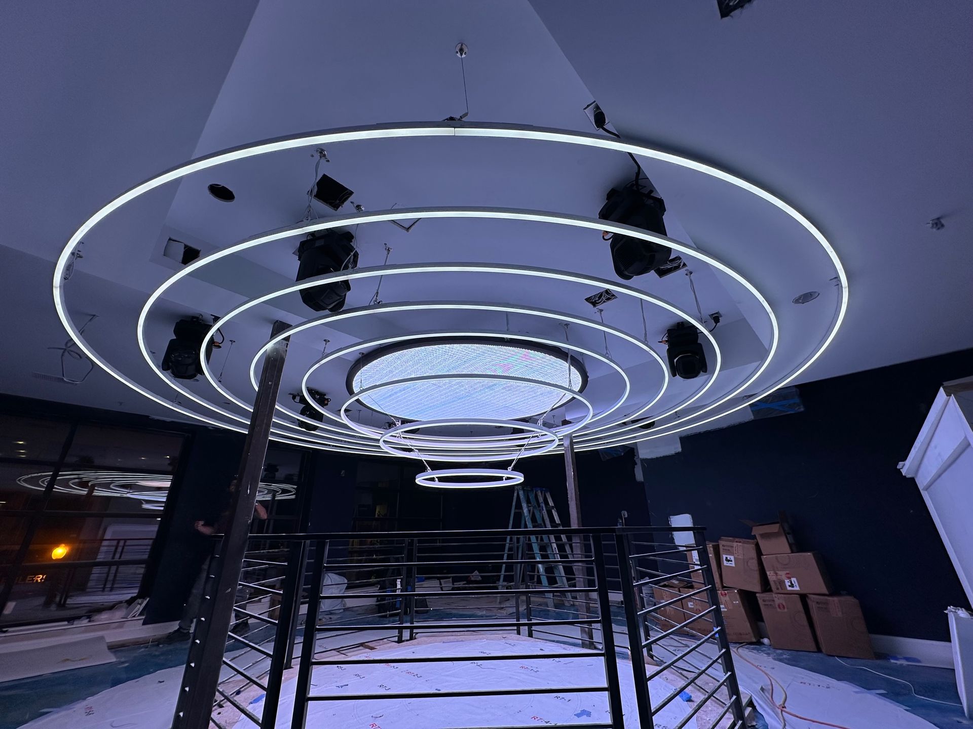 Circular neon lights illuminate a venue ceiling, with stage lights and stairs visible below.
