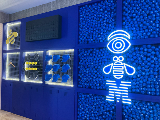 Blue wall with display niches containing yellow and blue shapes. A neon-light bee logo with an eye.