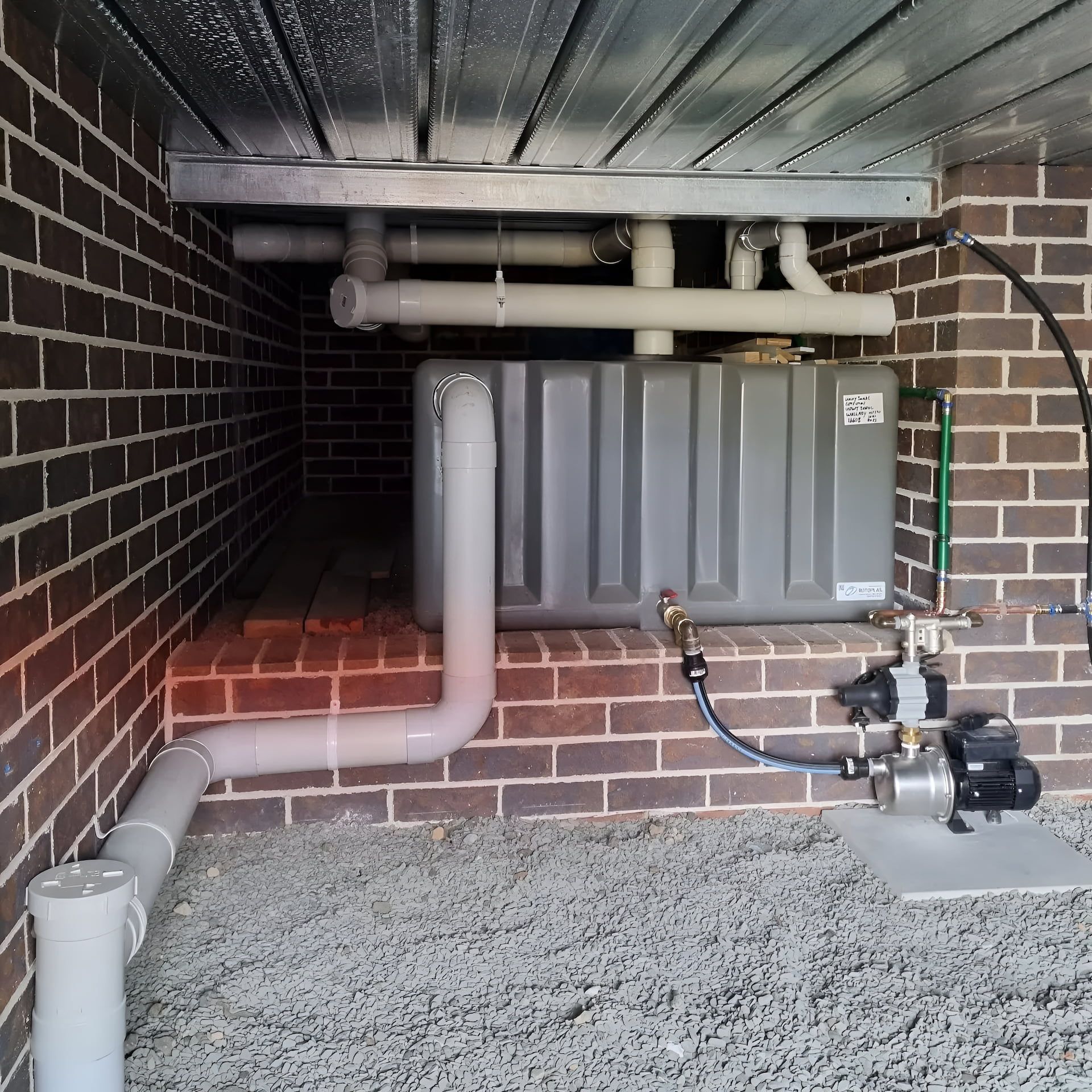 Water tank and pump system installed under a brick structure. Pipes connect to the tank and pump. — Corrimal Plumbing Services in Shellharbour, NSW