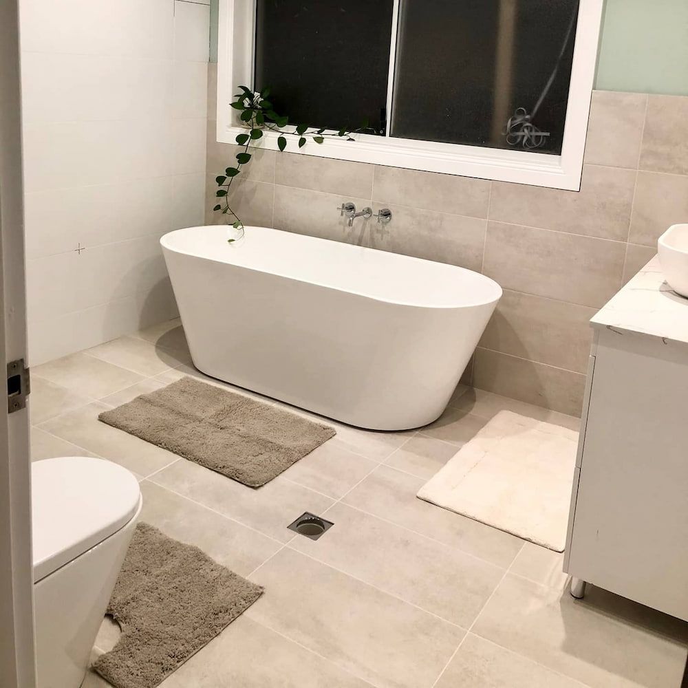 Modern Bathroom With a White Freestanding Bathtub, Two Rugs, and a White Vanity — Corrimal Plumbing Services in Bulli, NSW