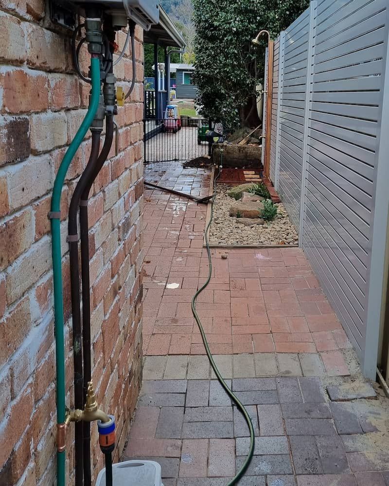 Brick Pathway Next to a Brick Wall With a Hose, Leading to a Fenced Area — Corrimal Plumbing Services in Thirroul, NSW