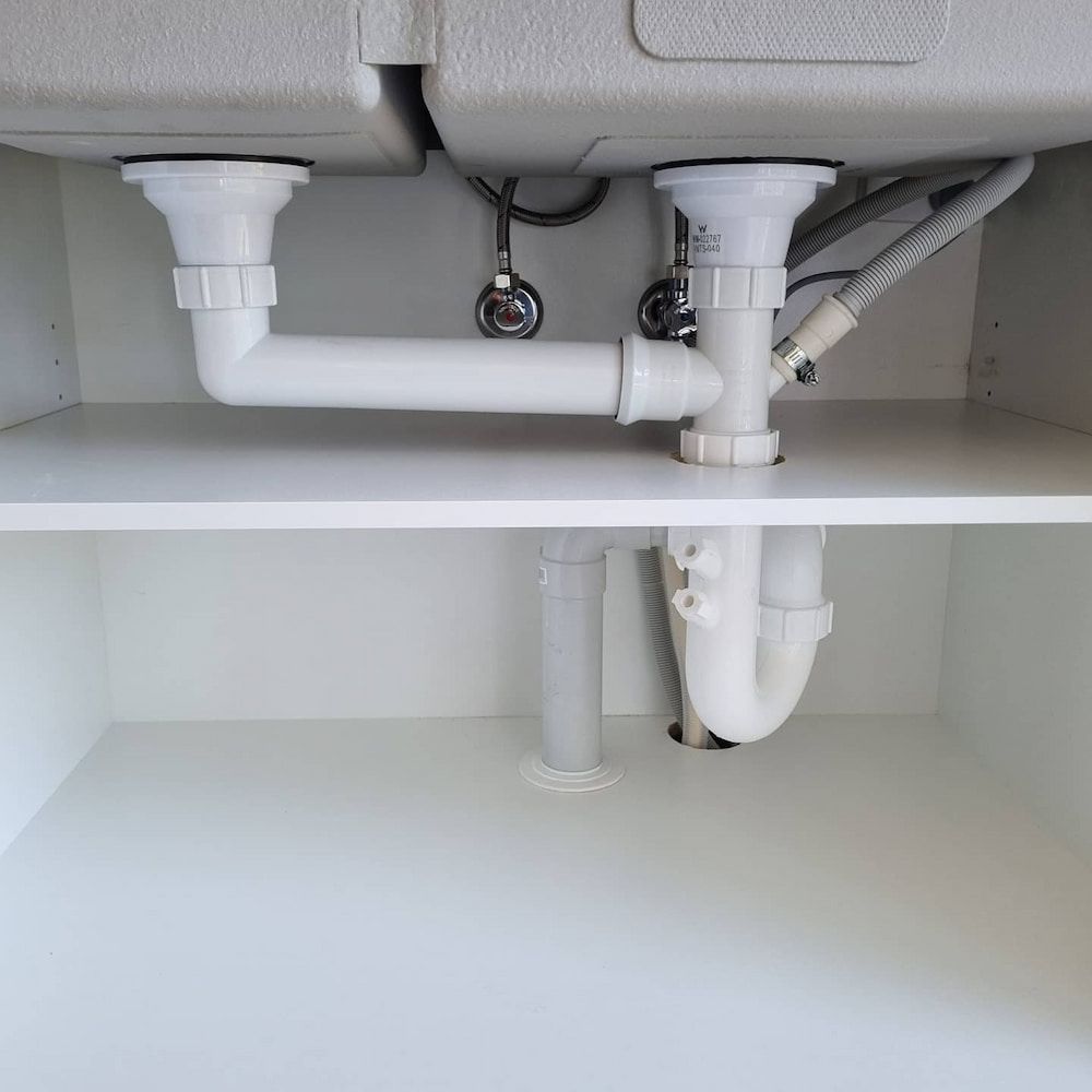 Under-sink Plumbing With White Pipes, a Double Basin, and Cabinet Interior — Corrimal Plumbing Services in Bulli, NSW