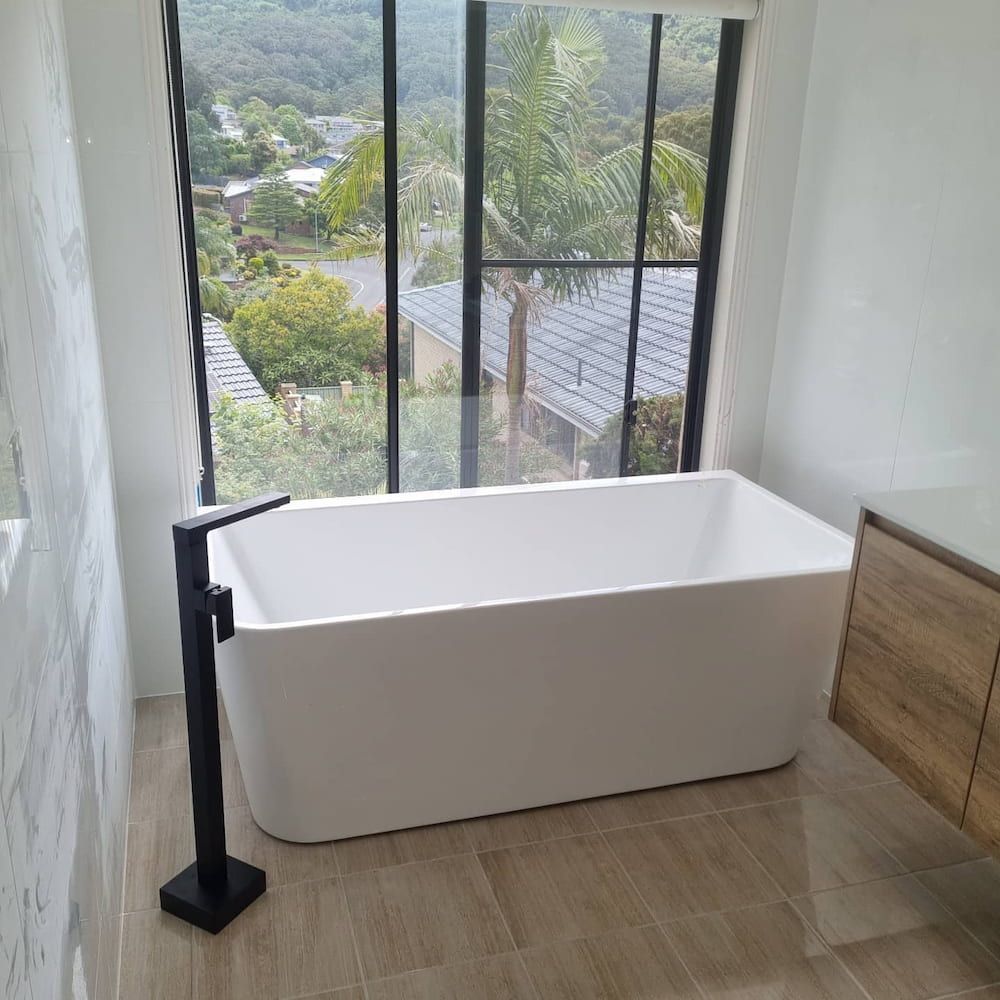 Modern White Bathtub Near a Large Window, Black Faucet and  Wooden Vanity — Corrimal Plumbing Services in Thirroul, NSW