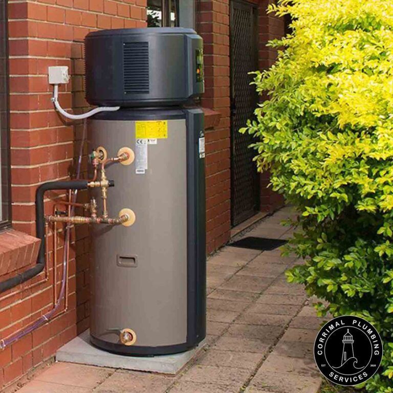 An Outdoor Heat Pump Water Heater Near a Brick Wall and a Bush — Corrimal Plumbing Services in East Corrimal, NSW