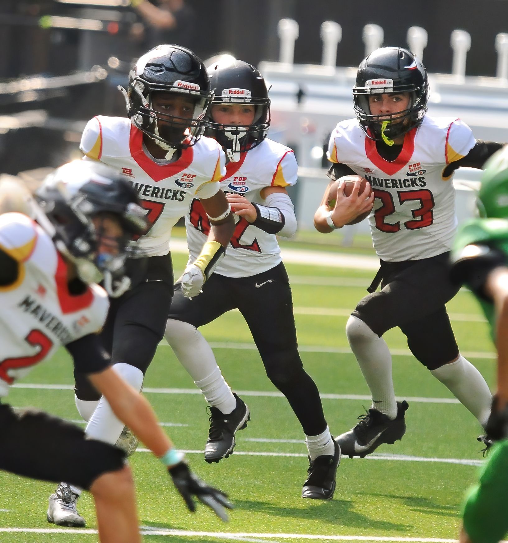 Football players in action on a grassy field; one runs with the ball, pursued by two opponents.