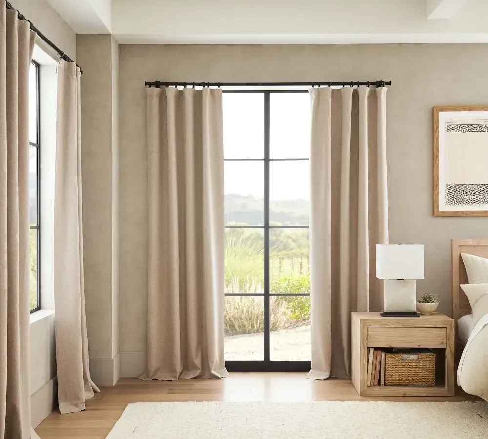 Bedroom with tan curtains, a large window, a nightstand, and a framed artwork.
