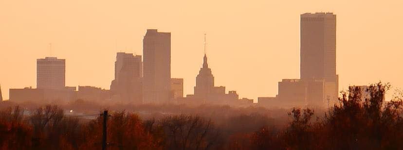 A city skyline is silhouetted against a sunset sky with trees in the foreground.