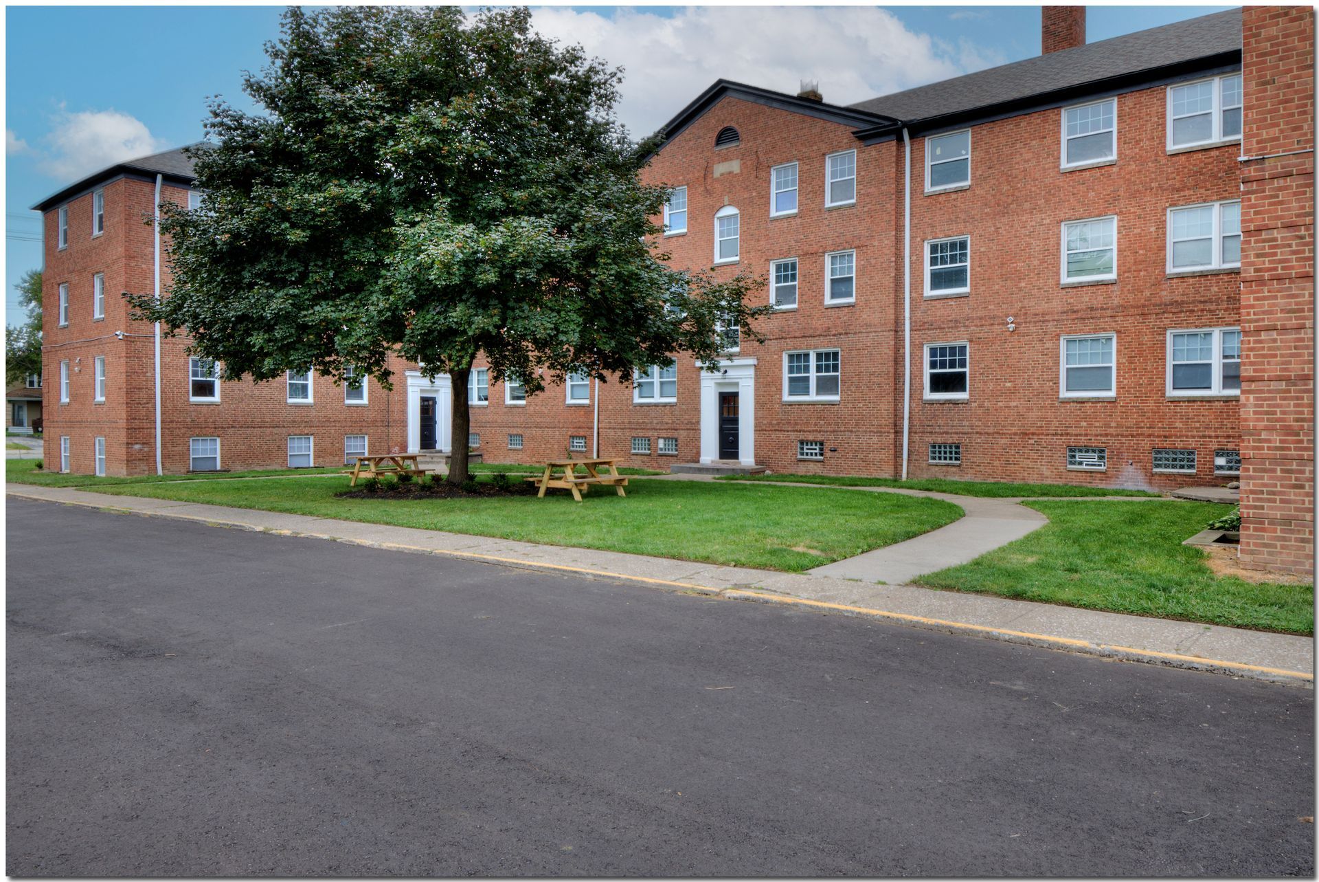 Red brick apartment building with a tree in front. Sidewalk, lawn, and paved street.