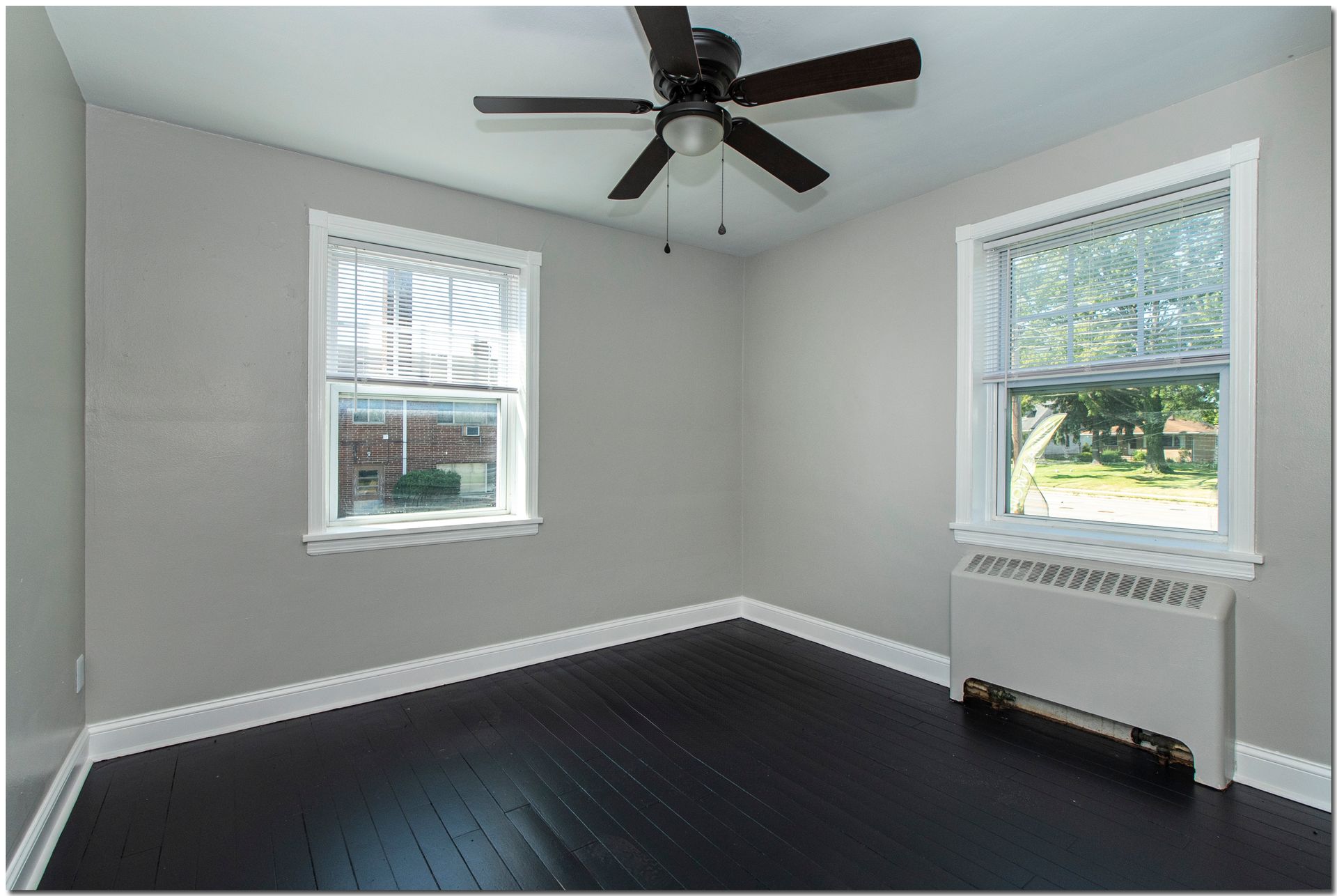 Empty bedroom with gray walls, dark wood floor, two windows, and ceiling fan.