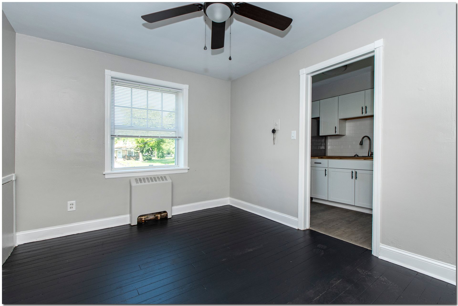 Empty room with dark floor, window, and doorway to a kitchen.