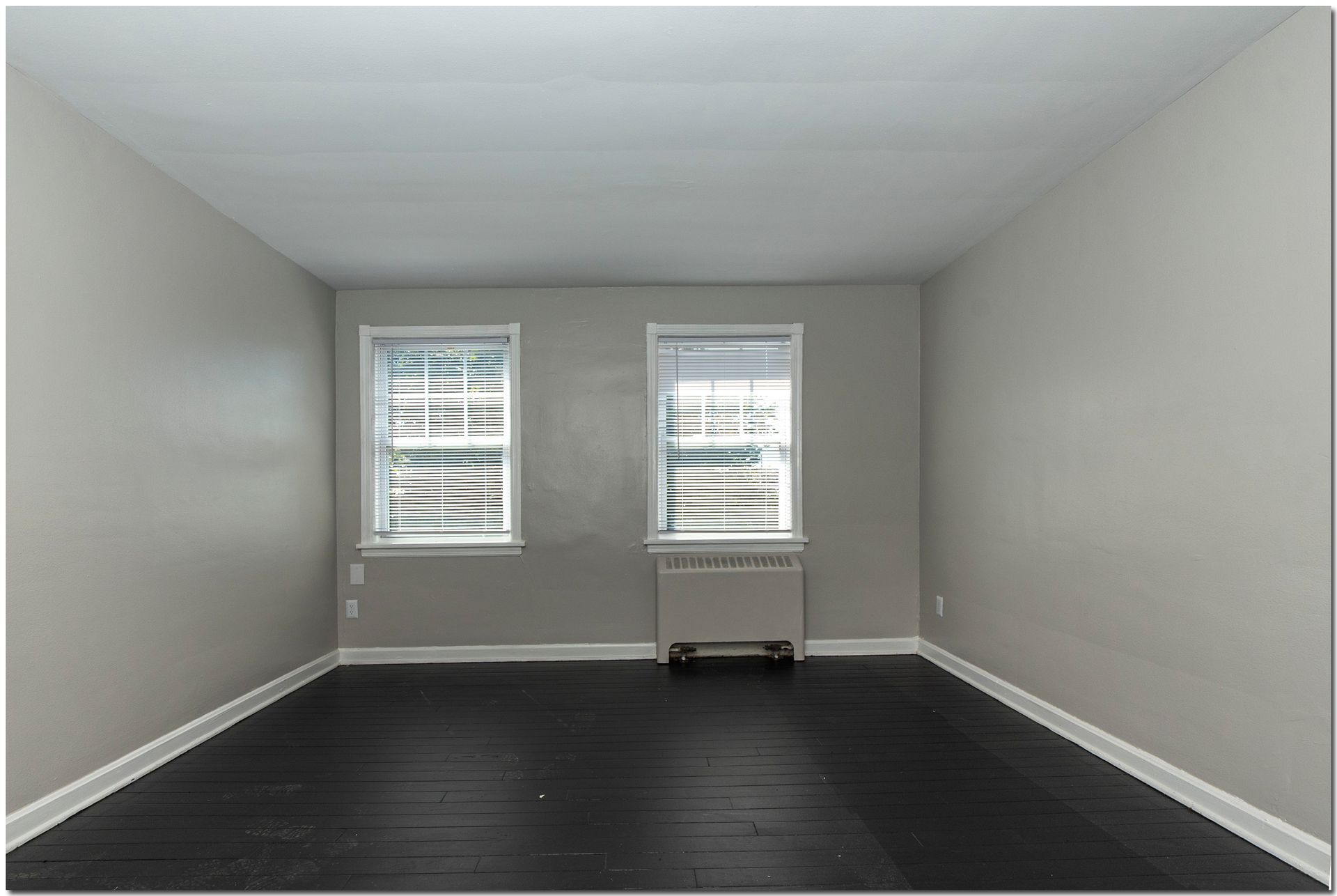 Empty room with gray walls, two windows with blinds, and dark wood floor.