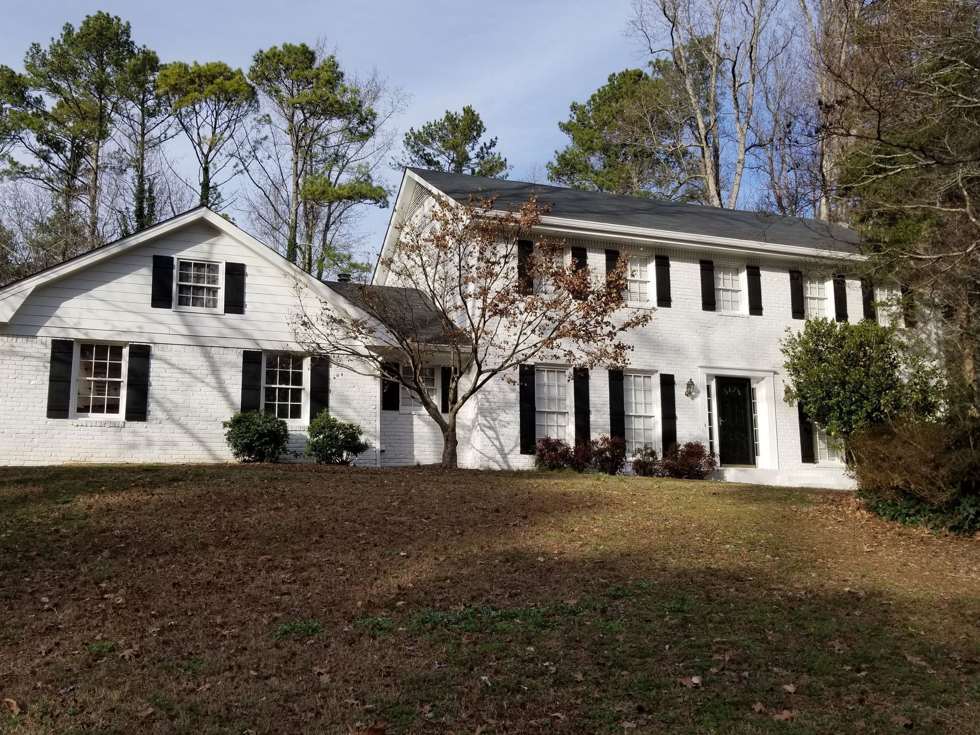 White two-story house with black shutters and door, set on a grassy hill with trees in the background.