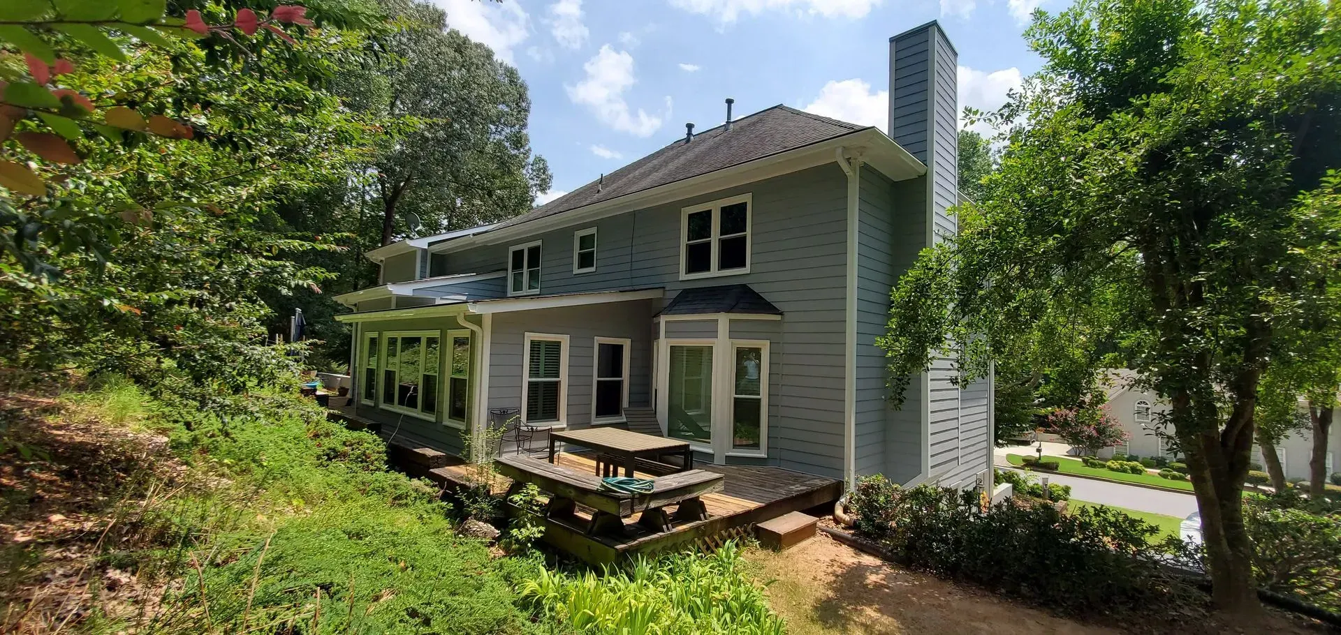 Back of a two-story gray house with a deck, surrounded by trees and foliage.